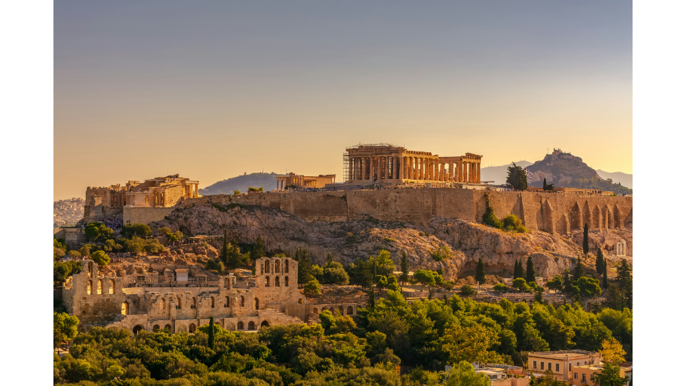 Sunset view of the Acropolis in Athens, Greece, with the Parthenon and surrounding ancient ruins standing above the city’s green hills.