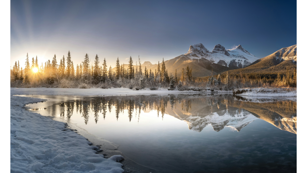 Snow-covered mountains reflected in a calm river with pine trees and sunrise light in the Canadian Rockies.