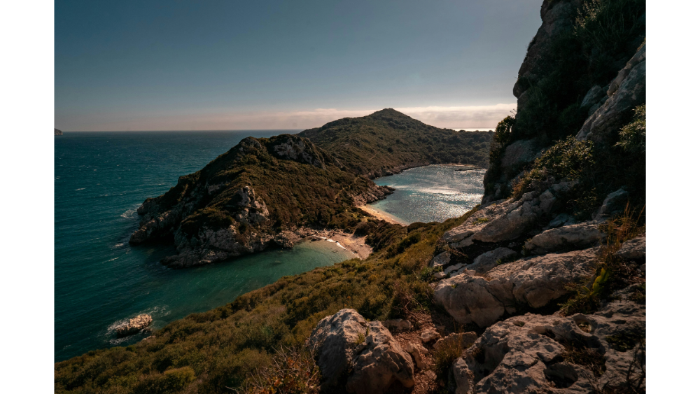 Scenic hiking view in Corfu with a rugged coastline, turquoise bay, and rocky hills covered in greenery under a warm, golden sky.