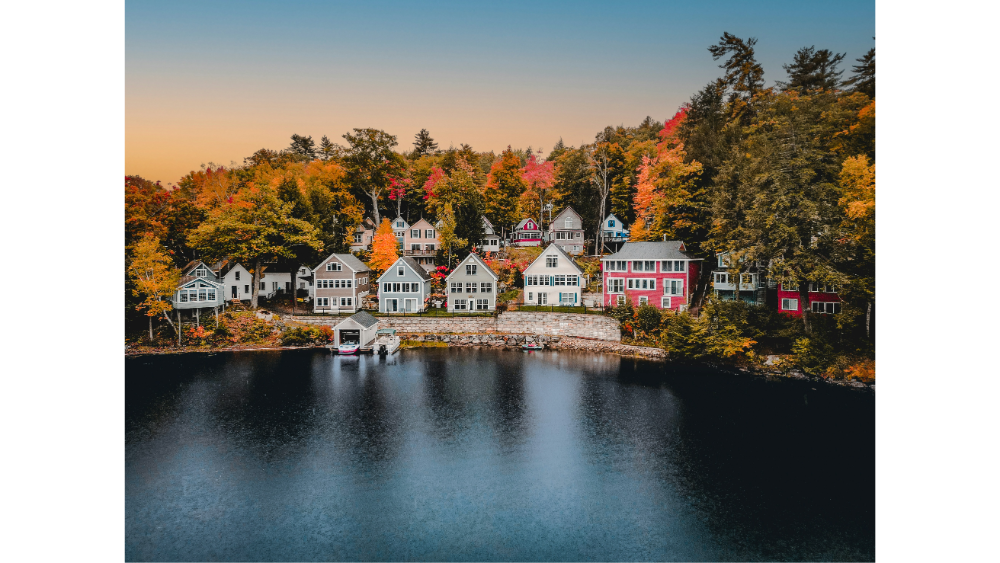 Lakeside village with colorful houses nestled among fall trees reflecting in calm water at sunset.