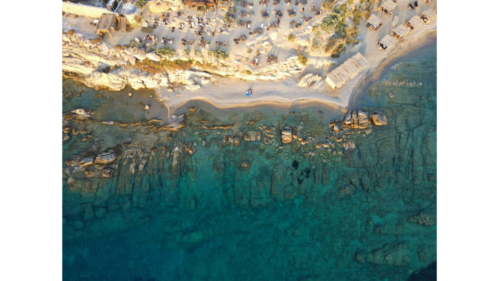 Aerial view of a rocky beach in Mykonos, with beachgoers, sun loungers, and crystal-clear turquoise water revealing the rocky seabed below.