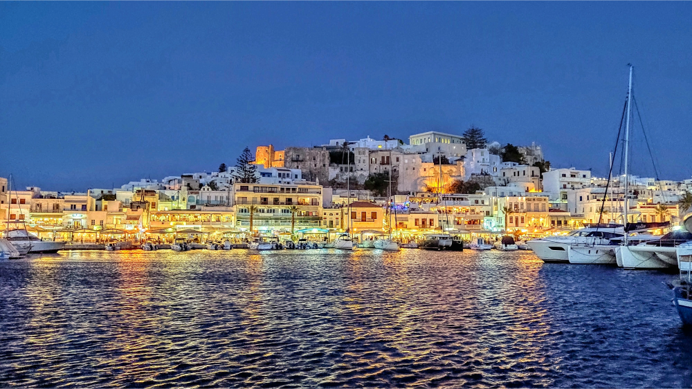 Night view of the Naxos harbor with boats docked and white Cycladic buildings lit up along the waterfront under a clear blue sky.