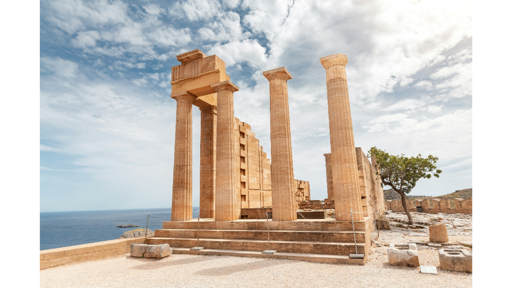 Ancient ruins of the Acropolis of Lindos in Rhodes, Greece, featuring tall stone columns overlooking the sea on a partly cloudy day.