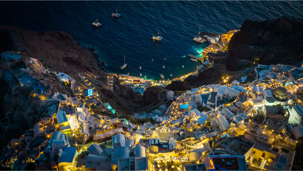 Aerial view of Santorini at night, with whitewashed buildings glowing under warm lights on the cliffside and boats floating in the deep blue Aegean Sea below.