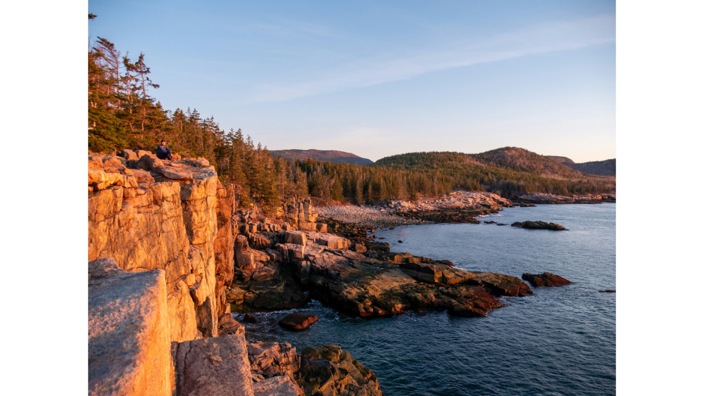 Sunset over rugged coastal cliffs and forested hills in Acadia National Park.