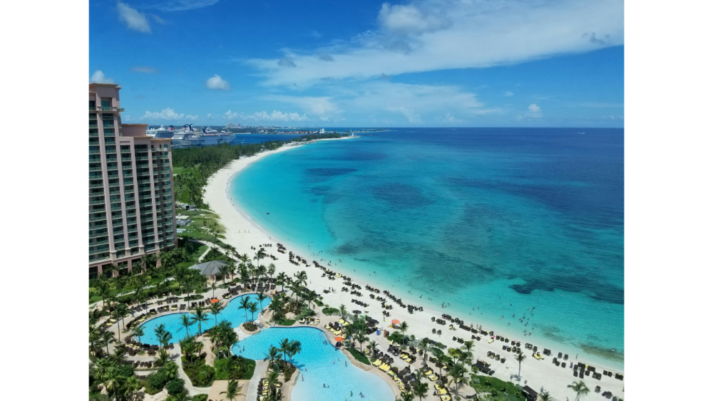 Aerial view of a beachfront resort with pools, lounge areas, and a curved shoreline next to turquoise ocean waters.