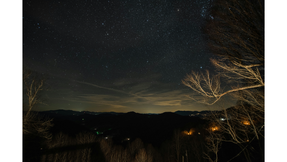 A nighttime mountain landscape with bare trees in the foreground and a star-filled sky above distant hills and scattered house lights.