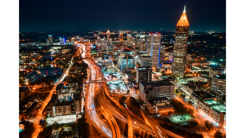 Aerial view of downtown Atlanta at night with illuminated skyscrapers and busy highways filled with light trails.