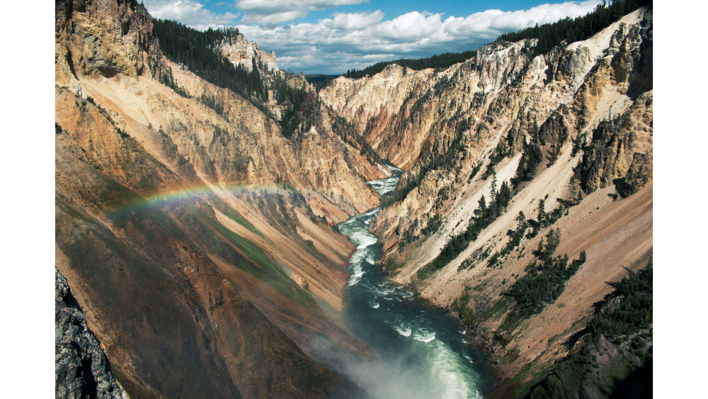 Steep canyon landscape with a winding river below and a vibrant rainbow stretching across the misty canyon walls.