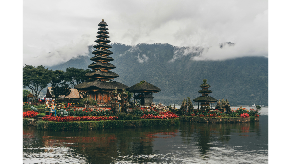 Traditional Balinese temple complex with tiered pagoda-style roofs, situated on a reflective lakeside with misty mountains in the background.