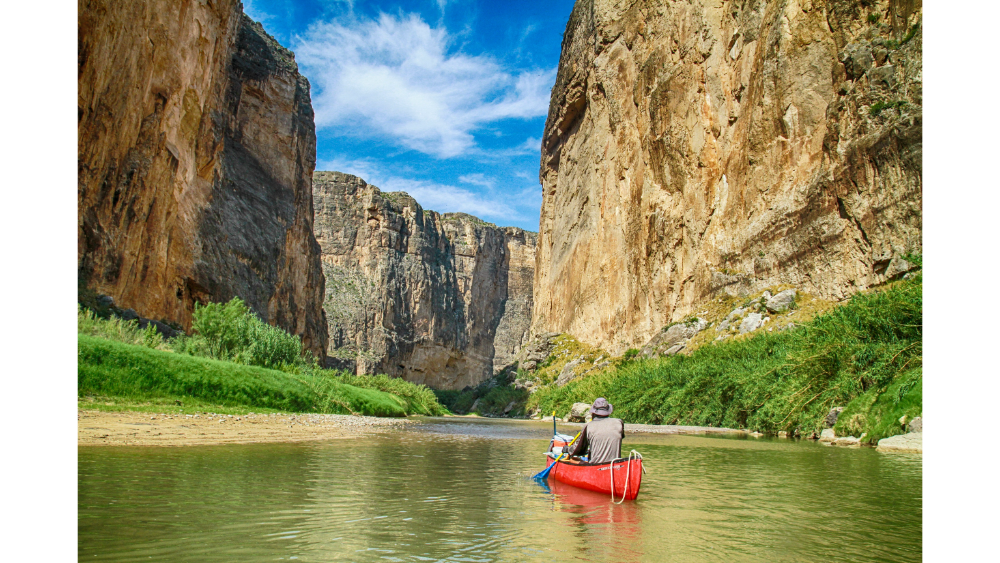 Two people in a red canoe paddling through a river flanked by tall, rugged canyon cliffs under a clear sky.