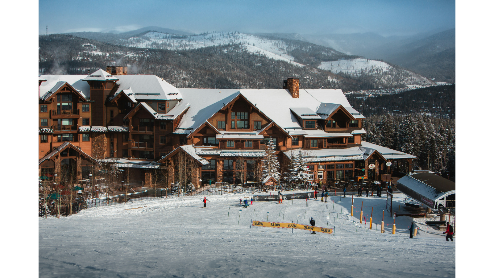 Lodge-style ski resort at the base of snow-covered mountains with skiers on the slope in front.