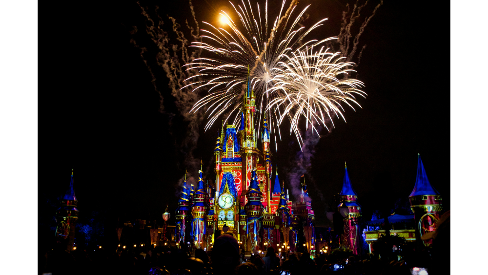 Colorful fireworks bursting above a brightly lit fantasy castle at night, surrounded by a crowd enjoying the show.