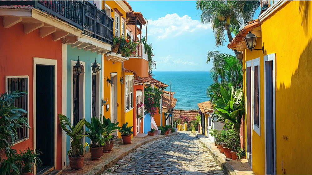 Cobbled street lined with vibrant colonial-style buildings and potted plants, leading down to a bright blue ocean horizon.