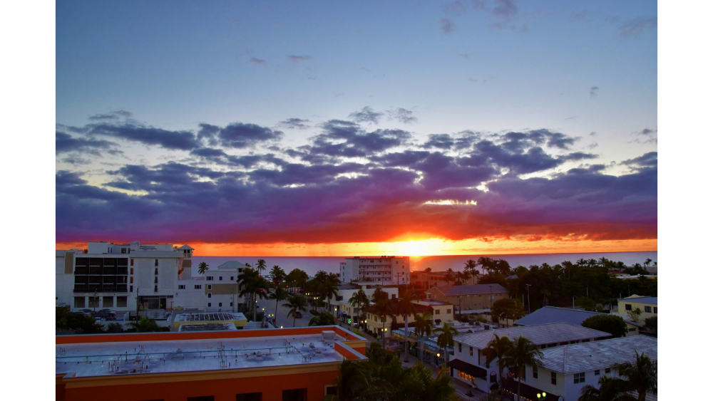 View of a coastal city skyline at sunset with vibrant orange and purple clouds above the ocean horizon.