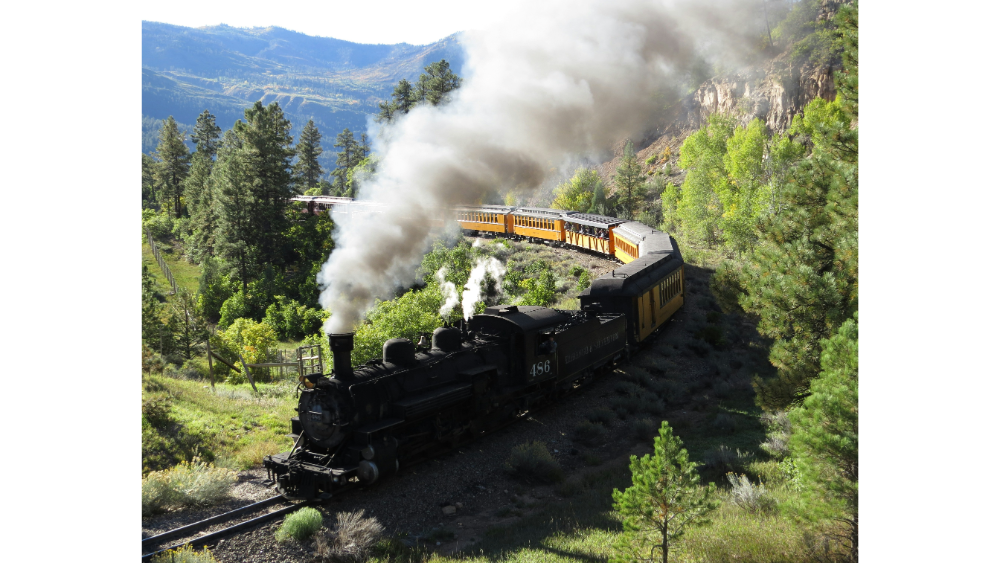 Vintage steam locomotive pulling yellow passenger cars through a forested mountain landscape.