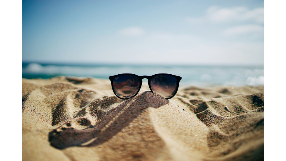 Pair of black sunglasses resting on sunlit beach sand with ocean waves softly blurred in the background.