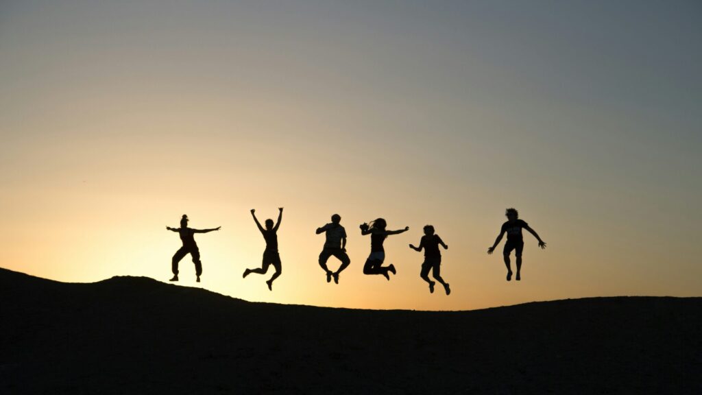 Silhouettes of six people jumping in the air on a hill during a colorful sunset.