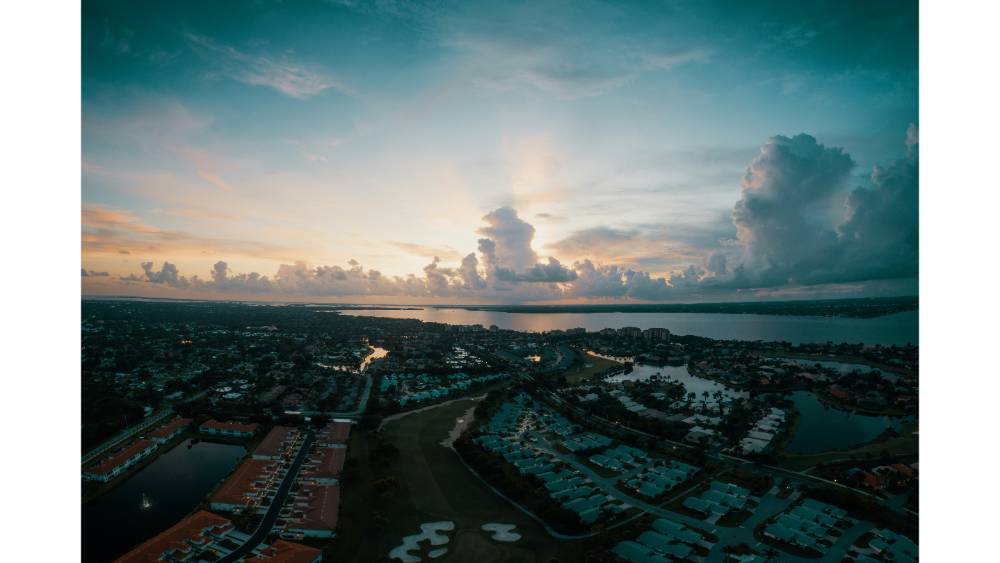 Sunset aerial view over a coastal city with houses, golf courses, and waterways under dramatic evening clouds.