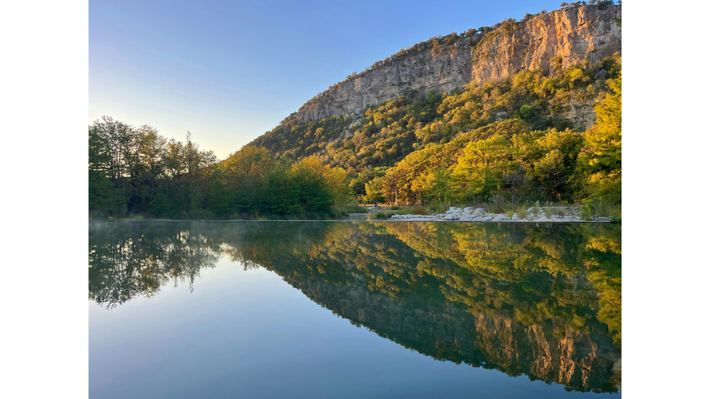 Peaceful landscape of Garner State Park with tree-lined cliffs reflecting perfectly in a calm, clear river during golden hour.