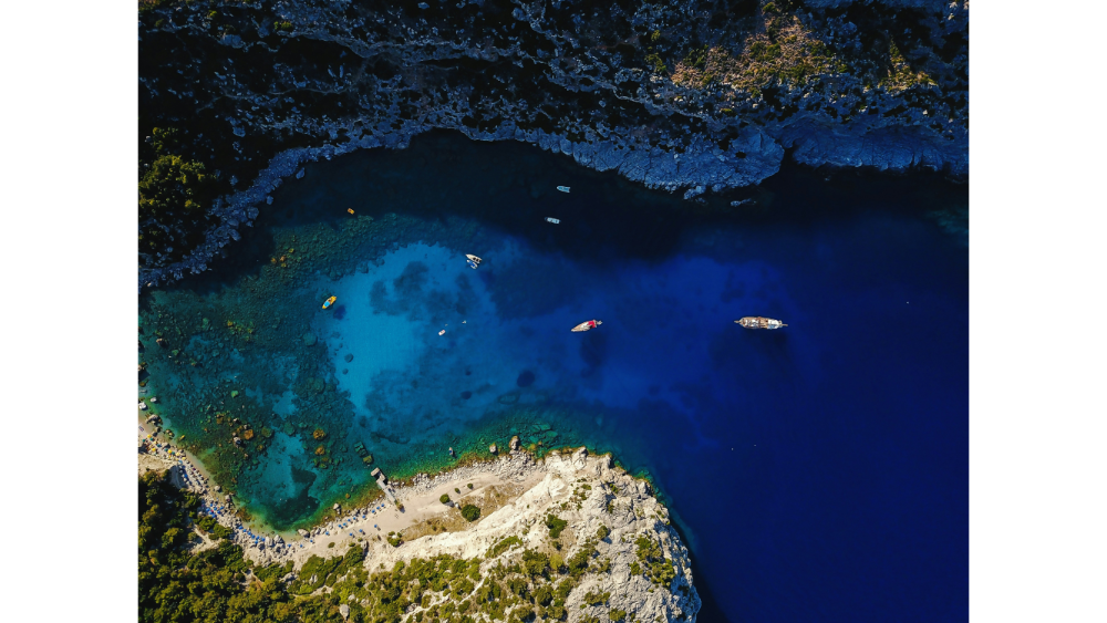 Drone view of a secluded tropical lagoon with vibrant blue water, anchored boats, and rocky shoreline surrounded by dense greenery.