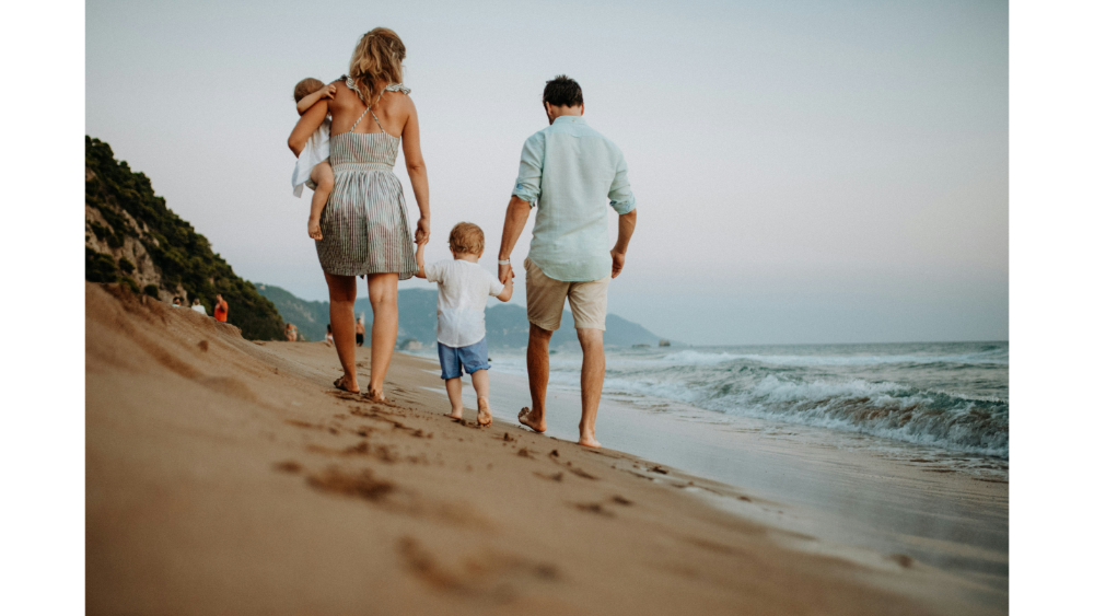 Family with two young children walking barefoot along a sandy beach at sunset, with gentle waves and distant mountains in view.