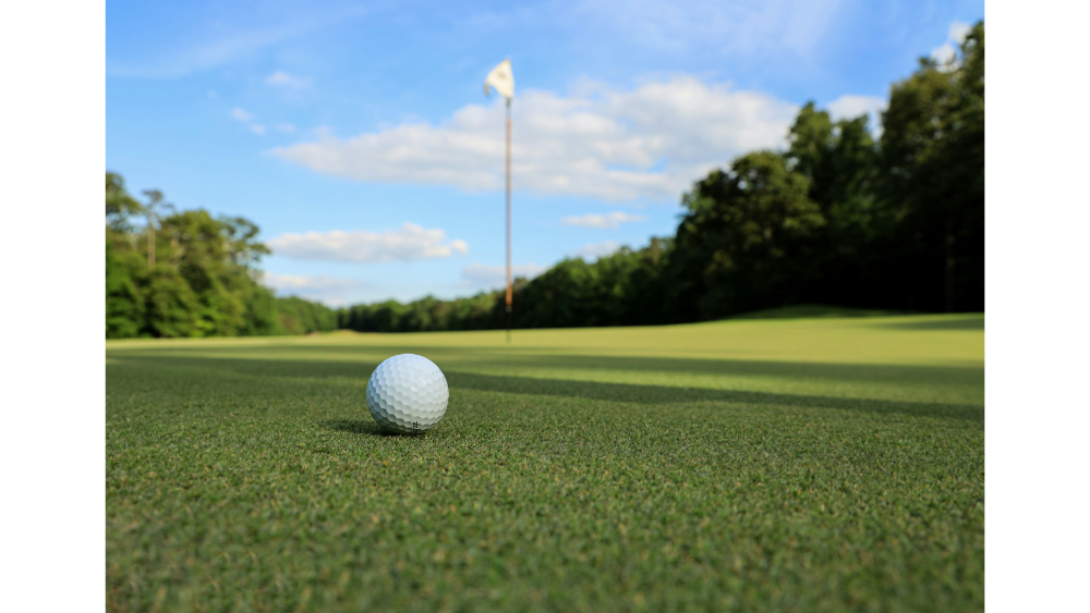 A close-up of a golf ball on the green with a flag in the distance under a clear blue sky.
