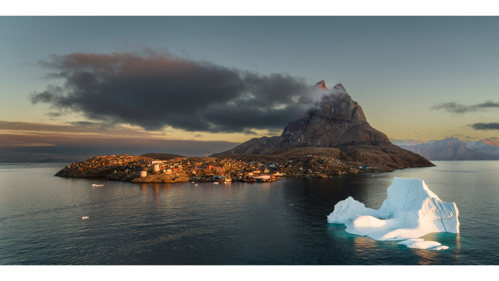 Large iceberg floating near a coastal village and towering rocky mountain under a dramatic Arctic sky.
