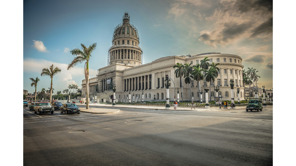 Historic capitol building in Havana, Cuba with vintage cars, palm trees, colonial architecture, and people walking along a sunny city street.