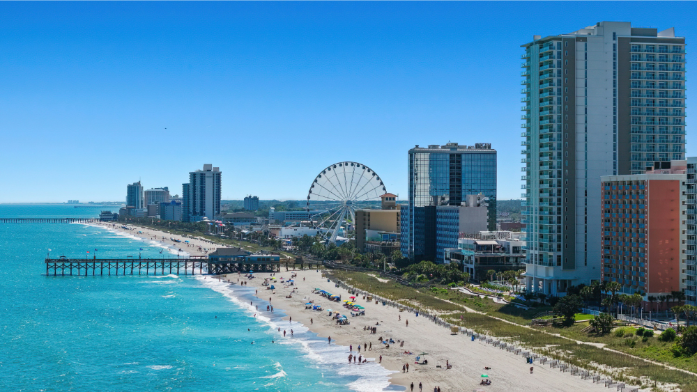 Coastal city with high-rise buildings, a Ferris wheel, and a long pier stretching into turquoise water along a sandy beach.