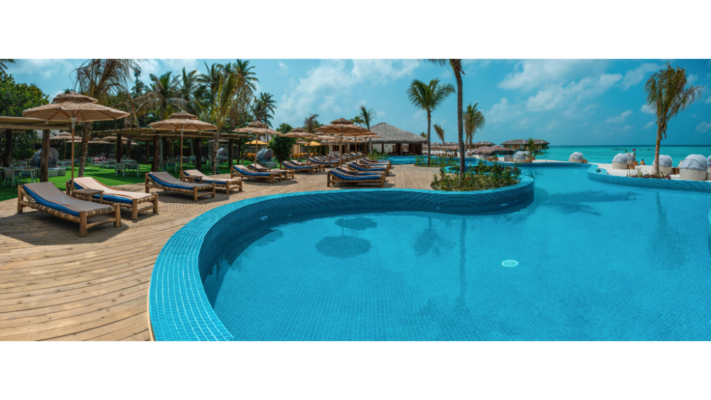 Large resort swimming pool with lounge chairs, palm trees, and a view of the ocean under a bright blue sky.