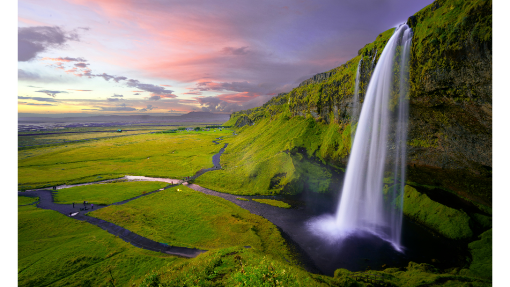 Tall waterfall cascading down a moss-covered cliff into a river winding through a vibrant green valley at sunset.