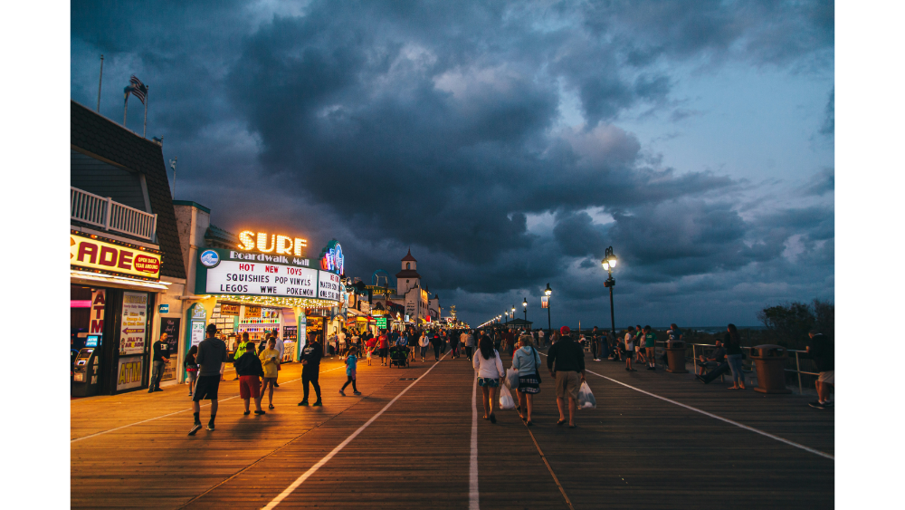 Lively boardwalk scene at dusk with glowing shop signs, people strolling, and dramatic storm clouds over the ocean.