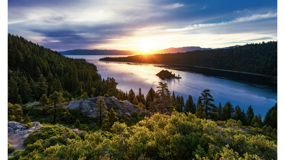 Golden sunset over Lake Tahoe with a forested shoreline, calm water, and a small island in the distance.
