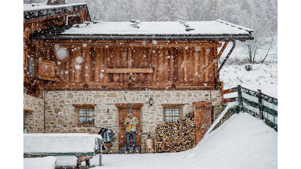 A rustic stone and wood cabin in a snowy landscape with heavy snowfall, stacked firewood, and a person standing by the door dressed in winter clothing.