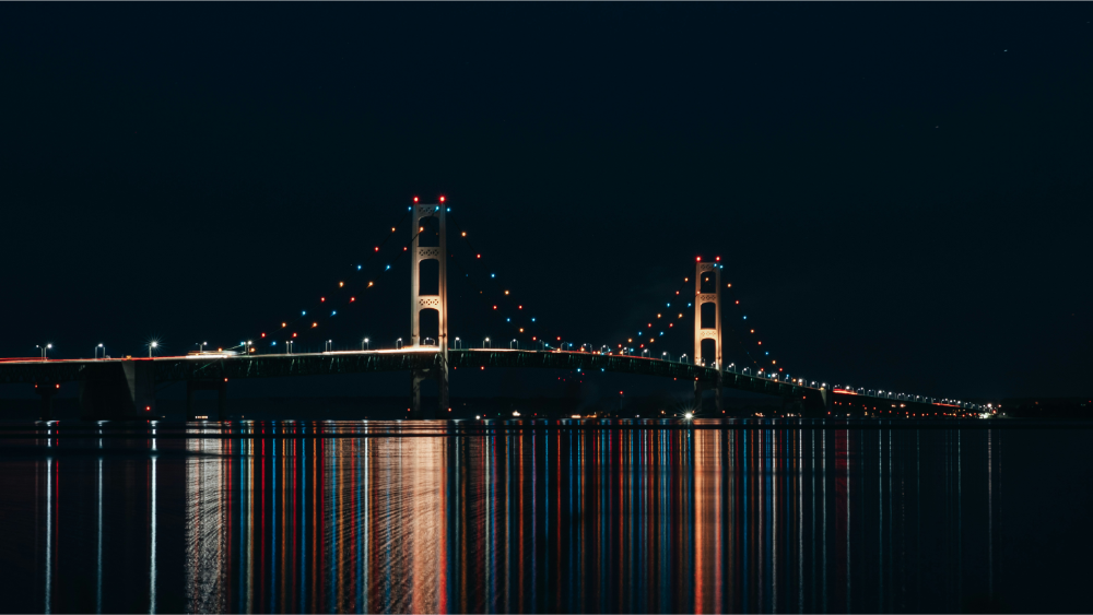 The Mackinac Bridge illuminated at night with colorful lights reflecting on the water below.