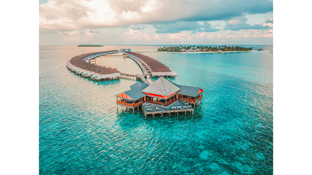 Curved row of overwater villas in a turquoise lagoon with a central resort building and nearby tropical island.
