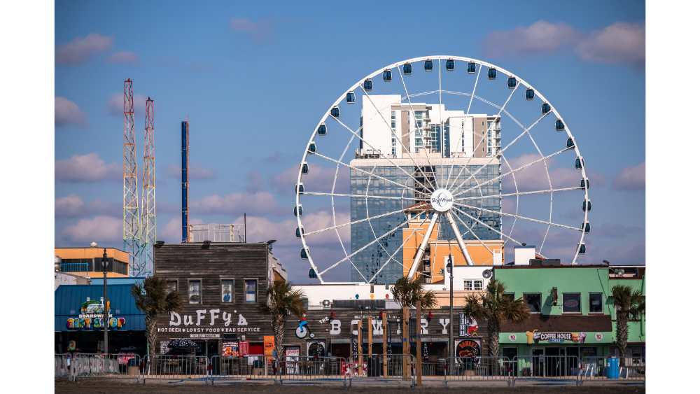 Large Ferris wheel rising behind a row of colorful boardwalk shops and restaurants with a cityscape in the background.