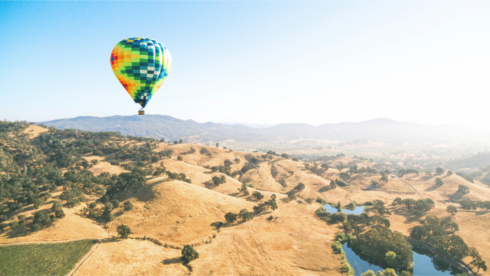 Brightly colored hot air balloon floating above golden rolling hills and vineyards with distant mountain views.