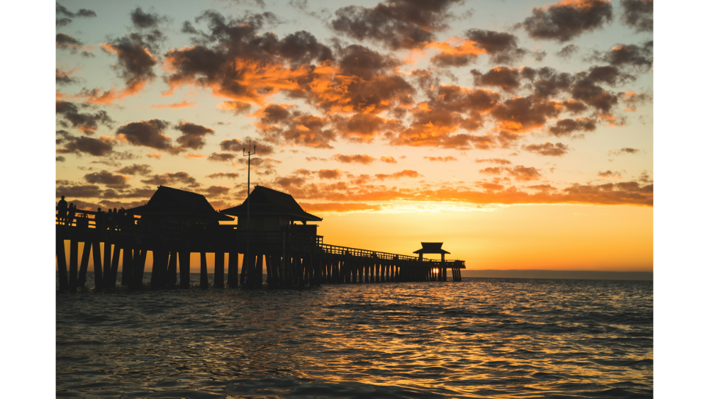Scenic ocean pier silhouetted against a vibrant orange and yellow sunset sky with scattered clouds.