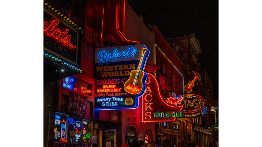 Bright neon signs for bars and music venues light up a lively city street at night, crowded with people.