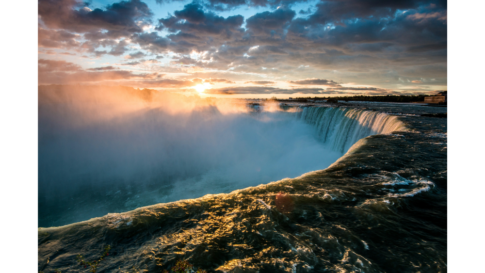 Majestic waterfall with mist rising as the sun sets in the background, casting golden light over the water at Niagara Falls.