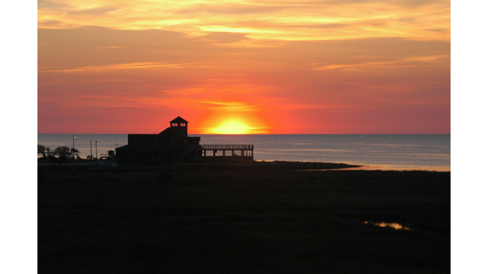 A vibrant sunset over the ocean with a silhouetted building in the foreground, located along a coastal landscape.