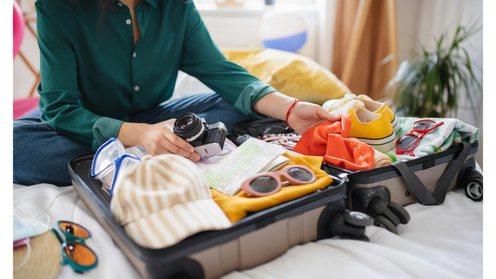 Person packing a suitcase with clothes, shoes, camera, and travel essentials on a bed.