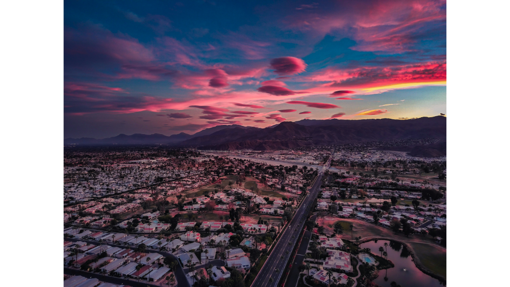 Aerial view of Palm Springs at sunset with dramatic pink clouds, desert landscape, residential neighborhoods, and surrounding mountain ranges.
