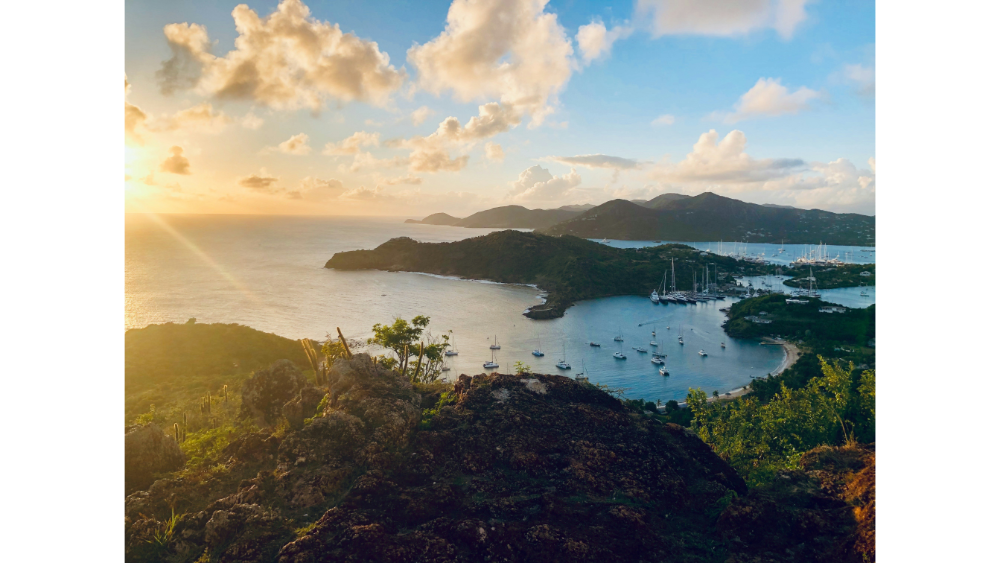 Scenic coastal view of a Caribbean bay at sunset with anchored sailboats, rocky cliffs, and lush green hills under a golden sky.