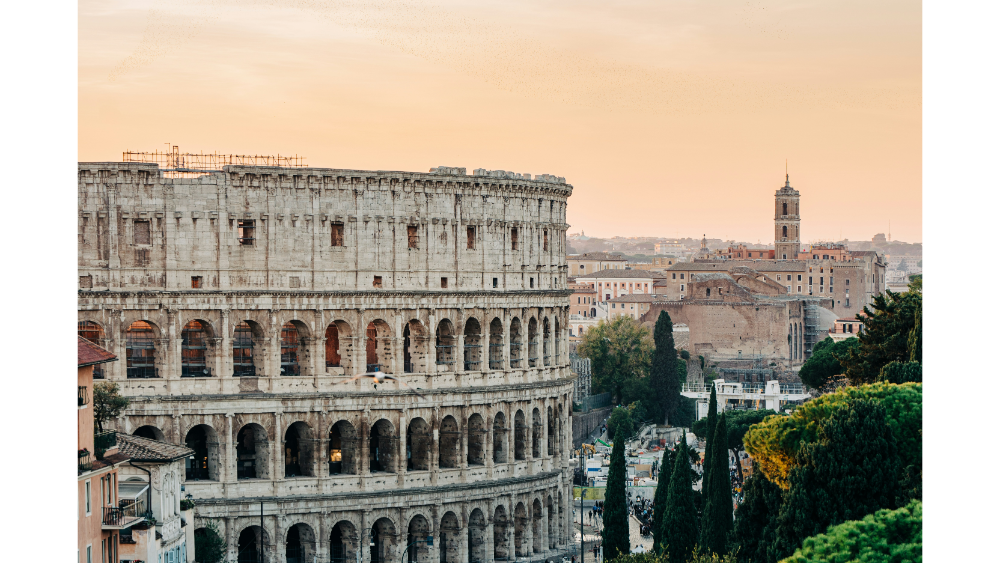Historic Roman Colosseum surrounded by ancient buildings at sunset with warm orange sky in the background.