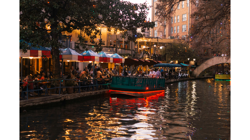 A festive evening scene at the San Antonio River Walk with diners, lights, and boats reflecting on the water.