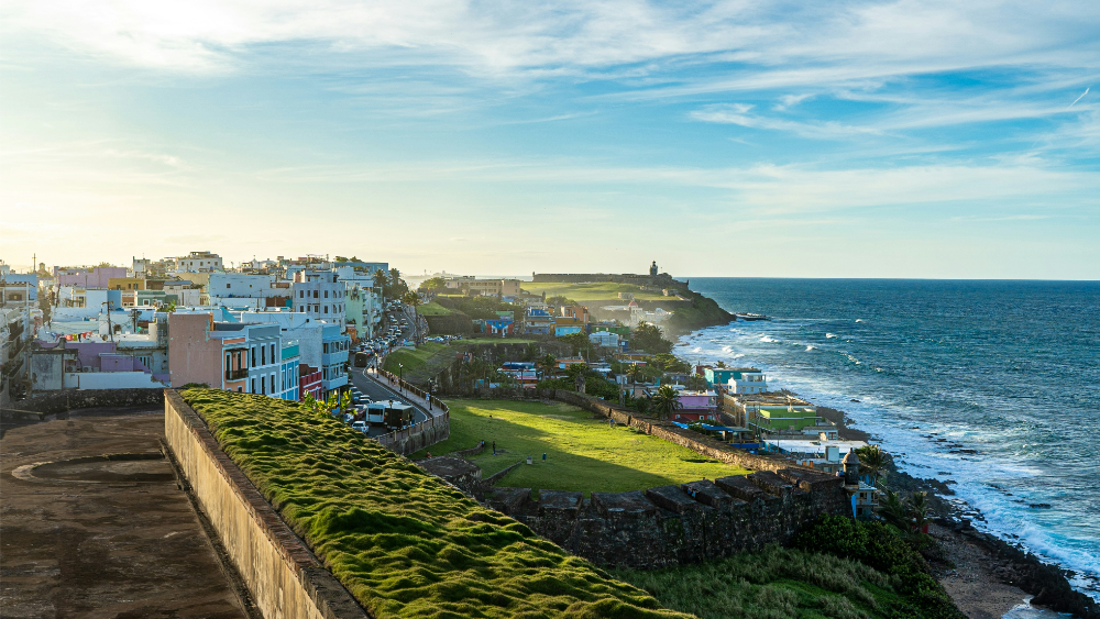 Scenic view of colorful buildings in San Juan, Puerto Rico, overlooking the ocean with historic stone walls, green lawns, and waves crashing along the rocky shoreline.