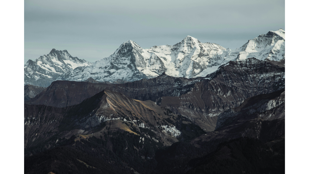 Dramatic view of rugged, snow-covered peaks rising above dark forested hills under an overcast sky.